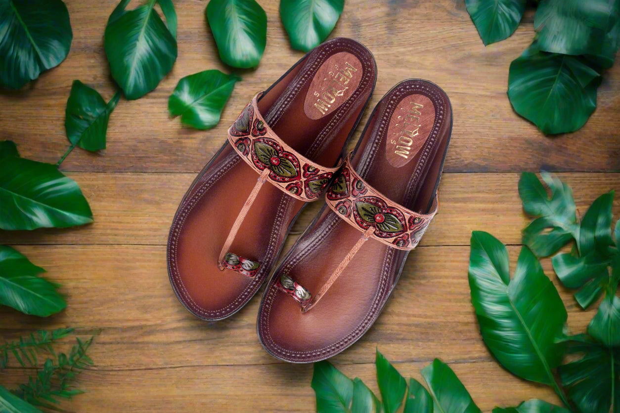 Pair of brown sandals with decorative straps on a wooden surface with green leaves.