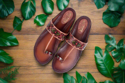 Pair of brown sandals with decorative straps on a wooden surface with green leaves.
