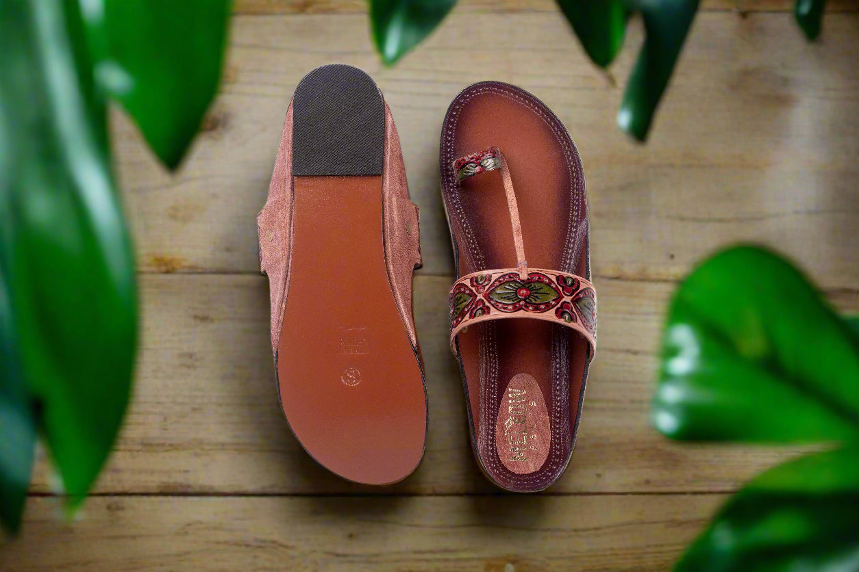 Pair of pink sandals with decorative straps on a wooden surface with green leaves.