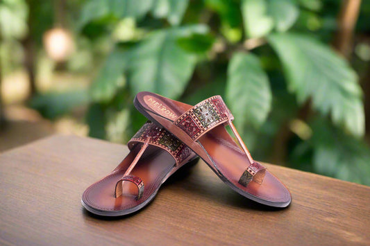 Pair of brown sandals with decorative straps on a wooden surface with a green leafy background
